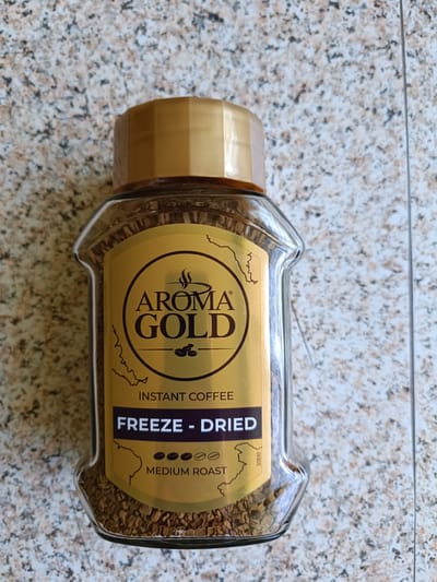 A clear glass jar filled with freeze-dried instant coffee granules is positioned vertically on a speckled granite countertop. The jar has a gold-colored screw-on lid. A prominent yellow label on the jar features the brand name "AROMA GOLD" in large, dark text. Below this, it states "INSTANT COFFEE" and "FREEZE - DRIED" in bold white letters on a dark purple background. Beneath that, five small brown circles indicate "MEDIUM ROAST". The background of the label is yellow with decorative brown outlines. The countertop is a light gray with irregular speckles of black and tan. The image is taken from a slightly elevated, overhead perspective. There are no people or animals present in the image.