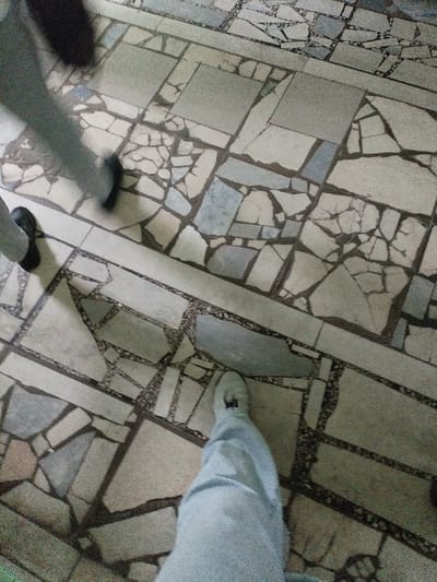 The image shows a view of a floor in Kalinkavichy, Belarus, located at latitude: 52.132, longitude: 29.347. The floor is made of irregular shaped pieces of gray and white stone, some of which are cracked. Two sets of feet and legs, belonging to two people, are walking across the floor. One person is wearing grey pants and black shoes. The other person is wearing blue jeans and a white shoe.