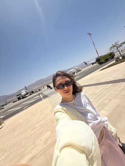 A person with dark hair and sunglasses is taking a selfie outdoors in Eilat, Israel. The person is wearing a white top and a pearl necklace. The background shows a paved area with stripes and a view of an airport runway with airplanes and a fire truck. Mountains are visible in the distance under a clear blue sky.