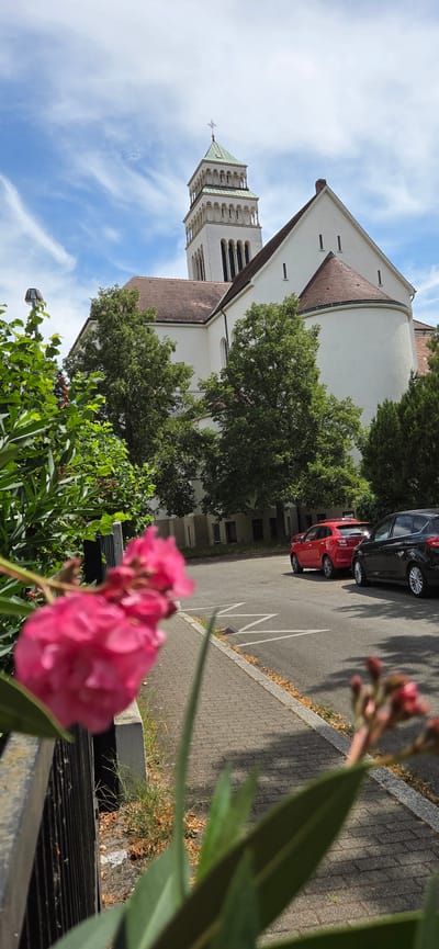 The image shows the church tower and facade of the St. Nikolaus Parish Church in Kehl, Germany. The church is a white building with a red-tiled roof and a tall, square tower topped with a green pyramidal roof and a cross. The tower has several levels with arched windows. The church is surrounded by lush green trees and foliage, with a pink flower prominently featured in the foreground, out of focus. In the background, there is a parking lot with a red car and a black car parked. The sky is blue with scattered white clouds. The scene appears to be a sunny day.