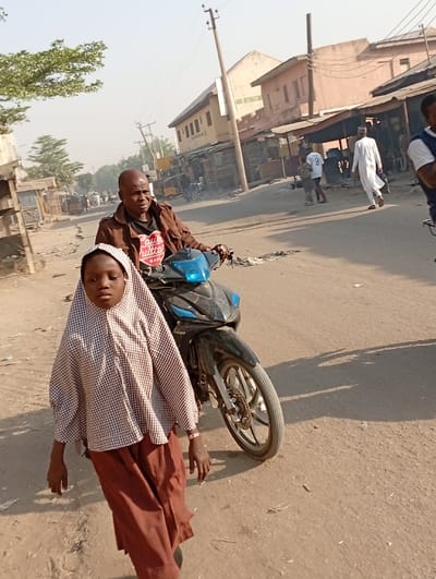 This image captures a street scene in Gama, Nigeria, featuring multiple individuals and structures under daylight conditions. In the immediate foreground, a young girl with dark skin walks forward, wearing a brown and white checkered long-sleeved top with a hood-like head covering and brown trousers; a light-colored bracelet is visible on her right wrist. Directly behind her, a bald, dark-skinned man in a brown jacket over a black t-shirt with a red 'Louis Vuitton' heart logo is positioned on a dark-colored LIFAN step-through motorcycle, his hands on the handlebars. The road surface is unpaved, dusty, and light brown. In the background, several one and two-story buildings line the street, some with corrugated metal roofs, suggesting commercial or residential use. Utility poles with overhead power lines are visible, crossing the frame. Other individuals are present further down the street, including a man in a white traditional robe walking from right to left, and another person wearing a white sports jersey with the number "9" on the back.