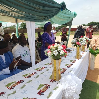 The image shows an outdoor gathering in Unguwar Saya, Nigeria. A group of people are seated under a tent with light blue and green drapes. A long table is covered with a white tablecloth adorned with a festive pattern of red trucks, Christmas trees, and snowflakes. Floral arrangements in vases are placed on the table.
Several individuals are visible in the foreground and background. A woman in a purple outfit and headscarf is looking towards the camera. A man in a white t-shirt with green text is also prominent. Other people are seated, some looking towards the table or engaged in conversation.
In the background, there are buildings, some appearing to be made of corrugated metal. Trees and a clear sky suggest it is daytime, possibly a sunny day. The overall atmosphere appears to be that of a community event or celebration.
Visible text on the t-shirt reads "MAJAY CA FOUNDATION".