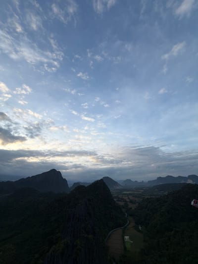 The image depicts a panoramic view from a high vantage point overlooking a valley in Ban Phon Sai, Laos, likely during sunrise or sunset. The sky is a canvas of pale blue and scattered white clouds, with warmer hues of yellow and orange peeking through on the horizon, suggesting early morning or late evening light.

Dominating the foreground are the dark, silhouetted shapes of rugged, tree-covered mountains and rock formations. The textures of these natural features are rough and jagged.

In the midground, a winding road is visible, cutting through lush green fields and patches of cultivated land, some of which appear to be rice paddies. A few hot air balloons are scattered in the distance, floating serenely in the sky, adding a touch of colorful vibrancy to the landscape. One balloon, in particular, appears to be red and white.

On the far right edge of the image, a portion of a hot air balloon or similar aerial apparatus is visible, featuring red and white checkered patterns. The overall impression is one of a serene, natural landscape, with hints of human activity in the form of agriculture and recreational ballooning. The lighting suggests a tranquil atmosphere, characteristic of a dawn or dusk sky.