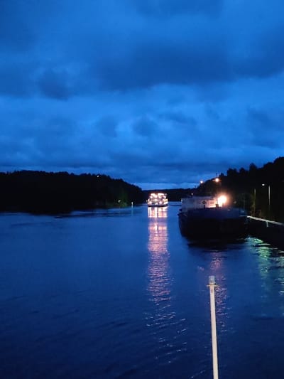 At dusk in Devyatiny, Russia, two cargo ships navigate a dark canal bordered by a dense, tree-covered shoreline. The sky is a deep blue, filled with ominous clouds. The water reflects the lights of the ships, creating shimmering trails. The lead ship is brightly lit, its windows glowing warmly, while the second ship, closer to the foreground, is partially obscured by darkness but also emits a warm light from its deck. The canal walls are visible on the right, with streetlights casting pools of illumination. A tall, slender pole with a light at its top stands in the foreground, its reflection stretching across the water. The scene conveys a sense of peaceful solitude, with the ships moving purposefully through the quiet evening.