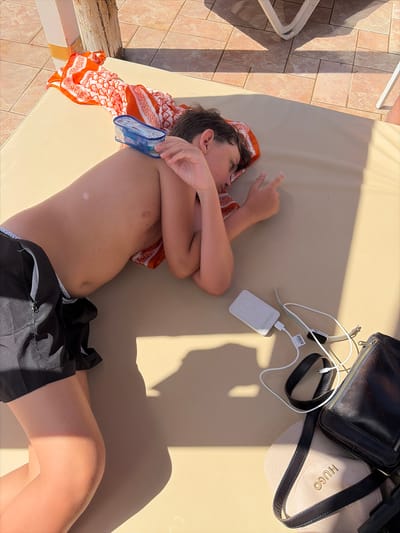 A young person is lying on a beige sun lounger, resting outdoors. The subject is wearing black swim shorts and has an orange and white patterned towel draped over their legs. To the right of the subject, a black leather bag, a beige cap with "HUGO" embroidered in gold, a white wireless charger, and a smartwatch with a white band are visible. The scene is illuminated by bright sunlight, casting strong shadows, suggesting it is a sunny day, likely midday or early afternoon. The background on the left shows a tiled patio with shadows from a light-colored structure. The overall impression is one of leisure and relaxation in a warm climate.