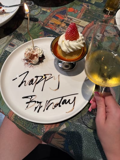 A dessert presentation is the focal point of a medium-angle shot, positioned on a patterned tablecloth and viewed from an elevated perspective. The dessert consists of a small ramekin filled with custard and topped with whipped cream and a strawberry slice, and a small mound of chocolate cake with whipped cream and cocoa powder. A silver spoon containing a lit birthday candle is placed near the chocolate cake. On the white plate, "Happy Birthday" is written in chocolate sauce. A hand holding a wine glass with a light-colored beverage is visible in the upper right corner, suggesting a toast. The setting appears to be a restaurant, indicated by the tablecloth and plate presentation. Zhangjiajie, China is not discernible from the details present in this image.