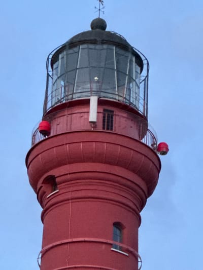 A close-up, low-angle shot shows the top portion of a bright red lighthouse, likely the Pakri Lighthouse, situated in Paldiski, Estonia. The lighthouse features a glass lantern room at its apex, topped by a dark dome and a weather vane. The lantern room is encased in a metal grate. Below the lantern room is a red, cylindrical section with a narrow walkway, accessible by a ladder. Two prominent red spherical lights are attached to the railing on opposite sides of this walkway. The lighthouse has a red, bulbous base with two small windows. The overall color palette consists of the vibrant red of the lighthouse against a muted, pale blue sky.
