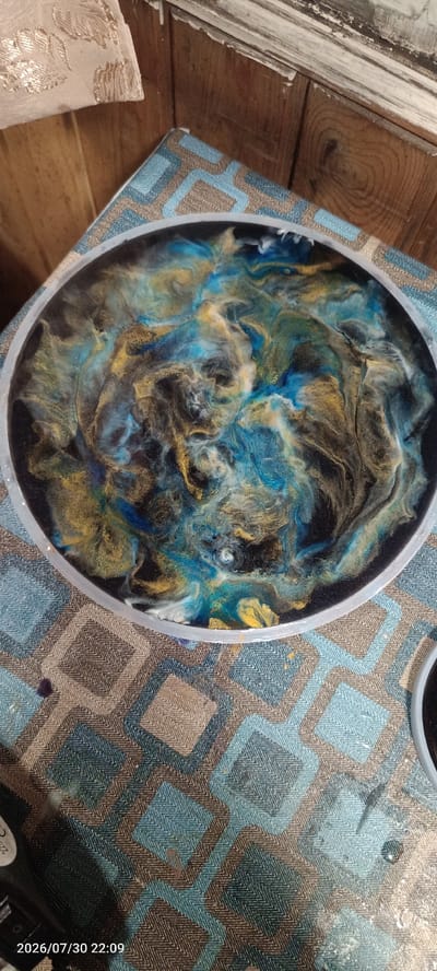 This image shows a close-up of a round, shallow mold filled with a resin art creation. The resin art features a swirling, abstract pattern of colors, predominantly deep blues, black, and metallic gold. The colors are mixed in a way that creates a sense of depth and movement, resembling a galaxy or a turbulent body of water. The mold itself has a clear, translucent edge, and it sits on a patterned fabric surface. The fabric has a geometric design with alternating squares of blue and brown, creating a chevron-like texture within each square.
In the background, there are elements of a wooden surface, possibly a table or wall, with visible wood grain. There's also a piece of decorative fabric with a floral or leaf pattern, possibly a tablecloth or tapestry, draped at the top left of the image. The overall lighting suggests an indoor setting, possibly during the day given the ambient light.
The activity taking place appears to be the creation or completion of resin art. The wet, glossy appearance of the resin indicates it is either freshly poured or still curing. There are no people visible in the image.
Visible text includes a timestamp at the bottom left corner of the image: "2026/07/30 22:09". There is also a partial label on an object at the bottom left, appearing to be part of a bottle or tool, with faint text that is not fully legible.
The setting appears to be a craft or workshop area, given the presence of resin art supplies and the materials used in the background. The location context of Severobaikalsk, Russia, suggests this is an activity taking place within that geographical area.