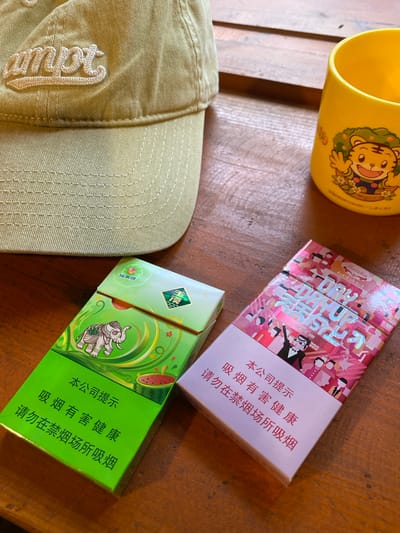 The image shows a close-up overhead view of a wooden table. On the table are several objects: a light green baseball cap with the word "ampt" embroidered in white, a yellow mug with a cartoon tiger illustration, a green pack of cigarettes with an elephant design, and a pink pack of cigarettes with a graphic pattern and text. The green cigarette pack has Chinese text that reads "本公司提示 吸烟有害健康 请勿在禁烟场所吸烟" which translates to "This company reminds you Smoking is harmful to health Please do not smoke in non-smoking areas". The pink cigarette pack also has Chinese text on it. The lighting suggests it is daytime. The image does not provide any specific information about the location within Dujiangyan City, China.