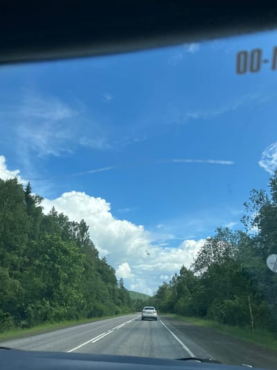 The image captures a view from inside a vehicle looking out at a two-lane asphalt road cutting through a lush, green forest. The sky above is a vibrant blue, adorned with fluffy white cumulus clouds and a faint, elongated cloud streak. Trees line both sides of the road, creating a tunnel-like effect. The road itself has white dashed lines marking the lanes, and a white car is visible ahead, traveling in the same direction. The scene appears to be set in a rural or natural environment, possibly on a highway or a main road through a forested area. There are no visible people or specific landmarks that would definitively place this scene in Усмангали, Russia, though the environment is consistent with many rural areas in Russia.