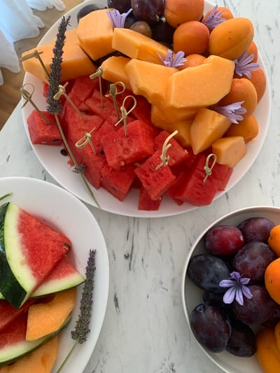 A high-angle, medium shot captures a close-up view of a fruit platter and a bowl of fruit arranged on a marble countertop. The platter, positioned centrally, is laden with cubed watermelon, cantaloupe slices, and apricots. Small decorative bamboo skewers, some with a knot at the end, are inserted into the fruit. Sprigs of lavender and small purple flowers are interspersed among the fruit, adding a decorative touch. To the left of the platter, a smaller white bowl contains slices of watermelon and cantaloupe, with a sprig of lavender placed alongside. To the right, a white bowl is filled with dark purple plums and apricots, with a delicate purple flower resting on one of the plums. The marble countertop has a white and grey marbled pattern. The scene appears to be indoors, likely in a domestic setting, possibly on a table near a window given the soft lighting. The location is not specified as being in Krimovice, Montenegro.