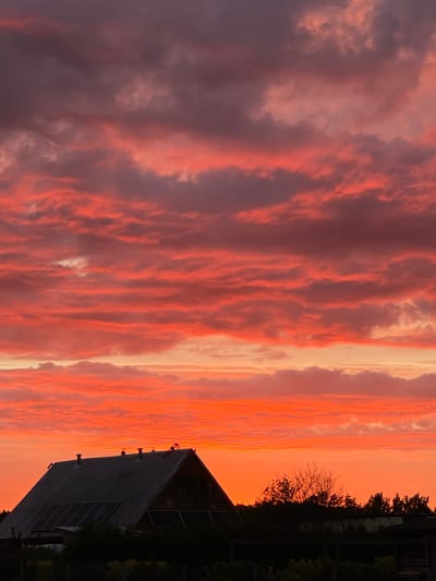 An eye-level, medium shot captures a vibrant sunset over a rural landscape in Zaķumuiža, Latvia. The sky is filled with a dramatic display of clouds painted in hues of deep red, bright orange, and purple, creating a striking contrast against the darkening silhouette of the land below. In the foreground, a dark roof of a house with several chimneys and solar panels is visible, suggesting a residential dwelling. A line of trees and vegetation forms a dark band across the bottom of the image, further emphasizing the silhouette effect. The overall atmosphere is one of natural beauty and tranquility.