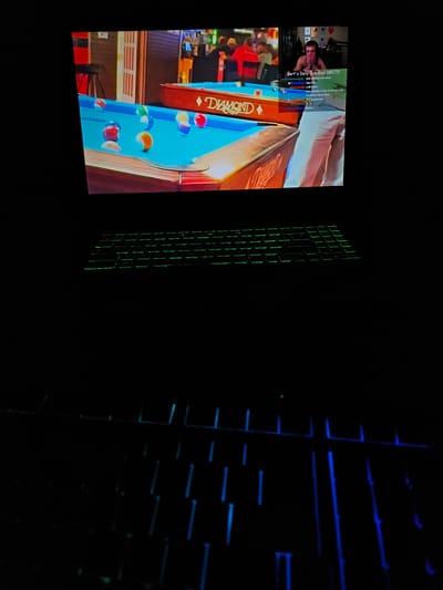The image captures a scene of a person playing a video game on a computer setup. In the foreground, a black keyboard with green-lit keys is visible. The background displays a computer monitor, showing a pool table game with several colored balls on a blue surface. There is also a small inset window in the top right corner of the monitor that appears to be a live stream feed, showing a person with headphones, looking stressed. Text overlays on the live stream indicate a "Bart's Daily Goal 188/75" and chat messages. The overall environment is dark, suggesting the image was taken at night or in a dimly lit room. There is no indication that this scene is located in Versailles, France.