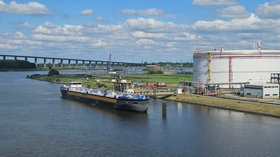 The image captures a wide-angle view of a river or canal in Brunsbüttel, Germany, on a clear day with scattered clouds. In the foreground, a long, dark-hulled barge named "RENATA REICH" is docked. The barge has a yellow stripe along its upper deck and a white cabin structure at the stern.

On the right bank of the waterway, a massive, white cylindrical storage tank with red accents dominates the scene. This tank appears to be part of an industrial complex, with numerous pipes and industrial structures surrounding its base. A small, grey building is situated near the tank.

In the background, a long, elevated bridge with numerous pillars spans the waterway, suggesting a significant transportation route. Beyond the bridge, wind turbines are visible on the horizon, indicating the presence of wind farms. The overall environment is industrial, with a mix of water, industrial infrastructure, and hints of green land and vegetation. There are no people visible in the image.