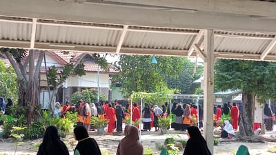 The image depicts an outdoor gathering of individuals, primarily women and girls, dressed in modest clothing, many wearing hijabs. The scene appears to be a school or community event, likely in Uteunreutoh, Indonesia, given the tropical vegetation and architectural style. In the foreground, a large tree with exposed roots is visible, with plants and potted greenery surrounding it. A white overhang structure, possibly part of a building or pavilion, frames the top of the image. In the mid-ground, a group of people are interacting, some standing and others appearing to be seated. The background shows buildings with tiled roofs, suggesting a residential or institutional area. The overall atmosphere suggests a communal event or gathering, possibly a religious or educational occasion.