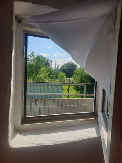 The image shows a view from an interior window, looking outwards. A white curtain billows on the right side of the frame, obscuring part of the view. Outside, lush green trees and a bright blue sky with scattered clouds are visible. In the middle ground, a corrugated metal fence runs horizontally across the scene, with another lower fence or railing in front of it. The window frame is simple and appears to be made of metal and white plastic. The interior windowsill is white and shows some signs of age and wear. There are no people or animals visible in the image. The scene suggests a residential setting, possibly a house or apartment building, in a location with abundant greenery.