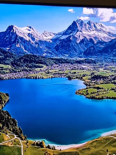 This is a high-angle, digitally rendered image depicting a serene mountain landscape with a lake and a village. The foreground features a vibrant blue lake with a light turquoise shoreline. Rolling green hills with winding white paths and scattered trees are visible on the right and lower left edges of the lake.
In the middle ground, a dense, green forested hillside slopes down to the lake. Nestled amongst the trees and on the lower slopes of the mountains is a small, picturesque village with buildings featuring light-colored roofs.
The background is dominated by majestic, snow-capped mountains. These mountains are rugged and jagged, with sharp peaks and dramatic textures. The sky above is a clear, bright blue with a few fluffy white clouds.
The image does not contain any people, actions, or interactions. The location in the city of Sambir, Ukraine is not depicted in this image, as it shows a natural mountain landscape.