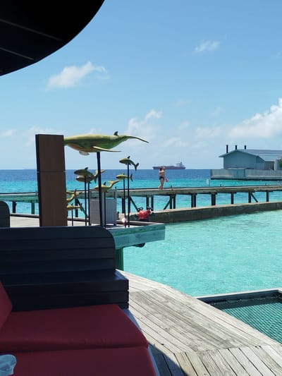 A woman in a bikini walks on a wooden pier over turquoise water in Gulhi Falhu, Maldives. She is carrying a red object. In the distance, a large ship sails on the horizon under a partly cloudy sky. A building with a grey roof and solar panels is visible on the right. On the pier, there are several large, green, fish-shaped sculptures mounted on poles, and a red fire extinguisher. In the foreground, a dark brown wooden bench and a red cushioned seating area are visible, part of an outdoor seating area. The water is clear and bright blue, with ripples on the surface.