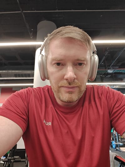 A fair-skinned adult male with light brown hair and a short beard faces the camera directly, wearing silver over-ear headphones and a red textured athletic t-shirt. The t-shirt displays a faint, mirrored "RUN" logo on the chest. He is positioned indoors, within what appears to be a modern gym or fitness center in Nahariya, Israel. The background features a dark ceiling with exposed piping and conduits, a prominent horizontal white light fixture, a large light-colored vertical support column, and blurred gym equipment, including weights and racks, visible in the lower portions of the frame.