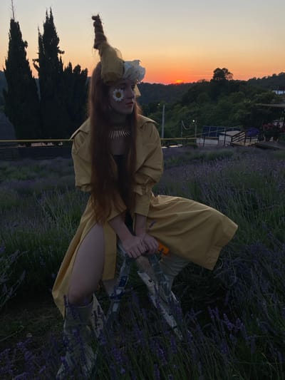A young woman with long red hair is sitting in a lavender field at sunset. She is wearing a yellow trench coat, a dark top, a metallic collar necklace, and tall light-colored boots. Her hair is styled with a large, elaborate accessory on top of her head. A daisy is attached to her left cheek, and white flowers are arranged near her eye. She is leaning on a wooden structure and holding an orange object in her right hand. The background shows rolling hills with trees, and the sky is a gradient of orange and pink from the setting sun. The location is identified as Verkhneye Uchdere, Russia.