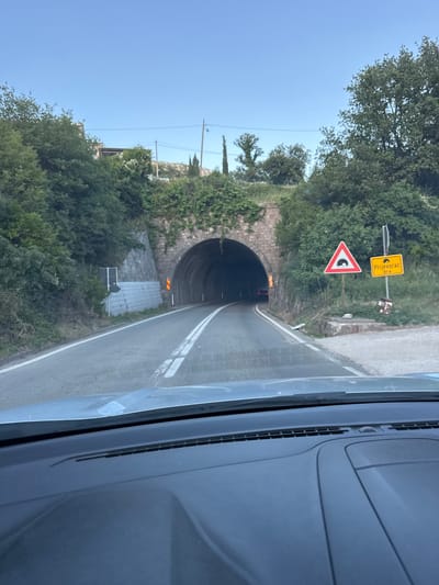 The image shows a forward-facing view from inside a vehicle, approaching a tunnel entrance. The road ahead is asphalt with white lane markings. A stone-arched tunnel is directly in front, with visible interior lighting and two red taillights of a vehicle further within. Green foliage and trees surround the tunnel and the hillside. On the right side of the road, there are two road signs. The triangular sign depicts a tunnel symbol. The rectangular yellow sign reads "Prijevorac 81m," indicating the name of the location and the distance to the tunnel. The sky is a clear blue with some distant, small specks that could be birds or insects. The foreground shows the interior dashboard of the vehicle. The scene appears to be in Prijevorac, Montenegro, given the road sign.