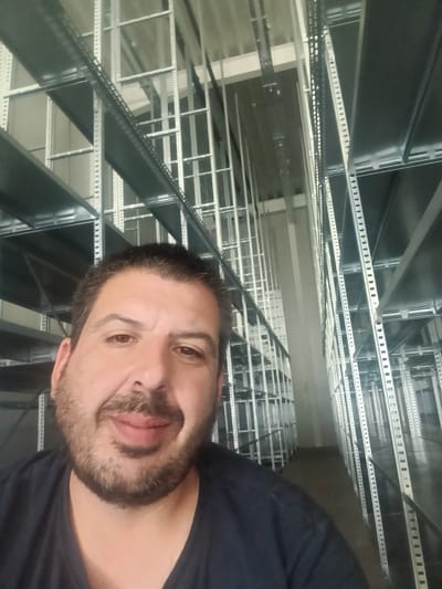 The image is a selfie taken by a man with dark hair and a beard, wearing a dark blue t-shirt. He is standing in what appears to be a warehouse or storage facility, with rows of tall, silver metal shelves stretching into the background. The shelves are empty. The lighting is somewhat dim, and the overall environment suggests an industrial setting. The location is identified as German, Bulgaria, but this detail cannot be visually verified from the image content alone.