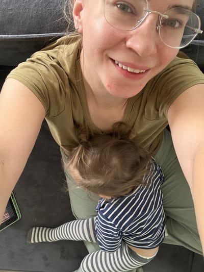 A woman wearing glasses and a green t-shirt is lying on a grey couch. A toddler is lying on top of her, wearing a blue and white striped shirt and grey and white striped leggings. A green tablet is visible in the lower right corner of the image. The location is unknown and the city of Kharnavy, Russia cannot be confirmed based on the provided image.
