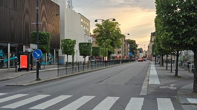 The image shows a street scene in Linköping, Sweden. On the left, a modern building with dark vertical cladding faces the street, with storefronts on the ground floor. A bus stop with a red illuminated advertisement is visible. A traffic sign indicates "Drottninggatan". The street itself is paved, with a pedestrian crossing marked by white stripes in the foreground. To the right of the street, a sidewalk is lined with trees and lampposts. Further down the street, there are more buildings, some with balconies, and shops. A few cars are driving on the road, and bicycles and scooters are parked on the sidewalk. The sky is overcast with hints of sunset colors. The overall impression is of a typical urban street during the day or early evening.