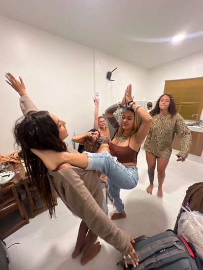 Five women pose in a row, with each woman stacked on top of the other, appearing to support each other. They extend their arms. The top woman wears a leopard-print outfit. The woman below her has brown hair and jeans and a brown top. The woman below her is blonde and wears a black top. The woman below her has curly hair and wears black. The woman in the front has long dark hair and wears a sweater. A router is visible on the wall.