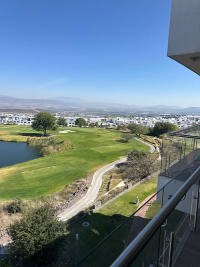 From an elevated perspective, a vibrant golf course unfolds with manicured green fairways and a winding cart path. A water hazard borders the course, adding to its aesthetic. White buildings dot the landscape, transitioning into a hazy mountain range in the distance under a clear blue sky. A glass balcony with metal railings frames the right side of the view.