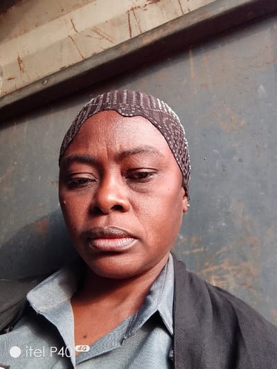 A woman faces the camera, wearing a patterned head covering. Her expression is neutral, and she appears to be indoors against a textured, grayish wall. She wears a blue collared shirt with a black jacket over it. A small gold earring is visible on her right ear. The lower left corner shows a logo.