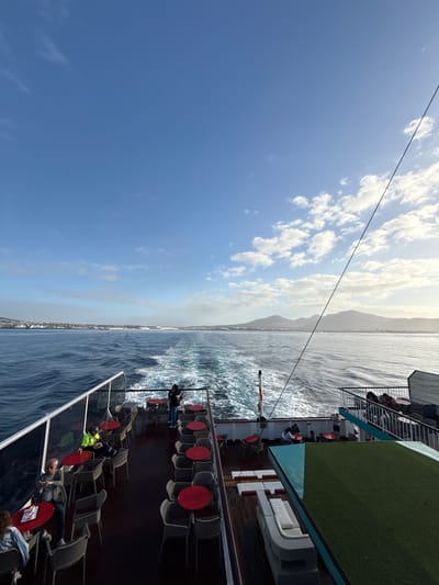 This image depicts a sunny daytime scene aboard a large ferry or passenger boat, looking out over the open sea towards a distant coastline. The boat is in motion, evidenced by the churning water and white caps created by its wake on the right side.

The sky, occupying the upper left of the frame, is a clear, vibrant blue with scattered white cumulus clouds. A dark cable or rope stretches diagonally across the upper portion of the sky. Along the horizon, a hazy mountain range or elevated landmass is faintly visible, suggesting an island or mainland coast in the distance.

The deck of the boat is prominent, featuring various seating areas. The lower deck has numerous grey, slatted chairs paired with circular red-topped tables. Several passengers are present, some seated, others standing, appearing to be relaxing and enjoying the journey. One individual is seen wearing a yellow high-visibility vest. Above this, a section of an upper deck is visible, covered in green artificial turf and furnished with modern white outdoor sofas. Metal railings separate the different deck levels and provide safety at the boat's edge.

The weather is bright and clear, indicating a pleasant travel day. The overall activity involves passengers traveling by sea, likely enjoying the outdoor views. No distinct, legible text is visible on any objects within the image. The setting aligns with a coastal or inter-island ferry journey, consistent with the location context of Yaiza, Spain.