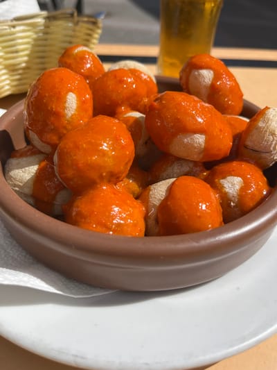 A bowl of papas arrugadas, a traditional Canarian dish, is shown on a white plate. The potatoes are small and wrinkled, covered in a bright red sauce. The bowl is brown ceramic, with the edges visible. The potatoes are arranged in a heap in the bowl. There are no people visible in the scene. The image was taken outdoors on a table, with a partial view of a glass of beer in the background. The location is in Agüimes, Spain, at coordinates 27.907, -15.447.