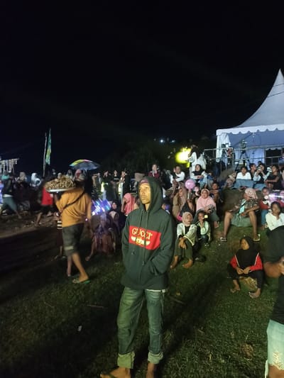 The image captures an outdoor night gathering in Kecamatan Pujut, Indonesia. The scene is dimly lit by artificial lights, with a large crowd of people spread across a grassy field.
In the immediate foreground, a young man stands facing slightly right, looking off into the distance with a neutral, possibly contemplative, expression. He is wearing a dark hooded sweatshirt with a prominent red stripe across the chest and the text "GUCCI" (stylized) visible. He also wears blue jeans and appears to be barefoot.
Behind him and extending into the mid-ground, numerous individuals, including adults and children, are seated or standing on the grass or on slightly elevated terrain, seemingly observing an activity or event. Many wear casual clothing, and several women are wearing headscarves. To the left, a person is seen walking while carrying a large tray, possibly with food items. In the mid-ground, a colorful umbrella is visible among the crowd, and a flagpole with a green and white flag stands further to the left.
On the right side of the image, a large white tent with a peaked roof is set up, possibly indicating a stage area or a vendor booth. Near the tent, a camera on a tripod suggests the event might be recorded or broadcast. A young person in the mid-ground holds a pink balloon. The overall atmosphere appears relaxed and communal, typical of a public event or festival taking place at night.