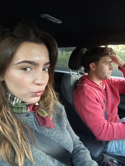 The image displays two individuals inside a vehicle. In the foreground, a female with light brown hair is visible, facing towards the viewer. She is wearing a grey textured garment and a multicolored striped scarf featuring green, white, brown, and maroon hues. Her lips are puckered, and a black seatbelt crosses her chest. Behind and to her right, a male subject with light brown hair is seated, looking towards the right side of the vehicle, out of frame. He is wearing a red hooded sweatshirt, secured by a black seatbelt. His right hand is placed on his forehead. The vehicle's interior features dark upholstery and a dark headliner. Through the windows, blurred green foliage and ambient light are discernible, indicating an outdoor setting during daylight. The specific location within the city of Leiria, Portugal, cannot be determined from the visual evidence provided.