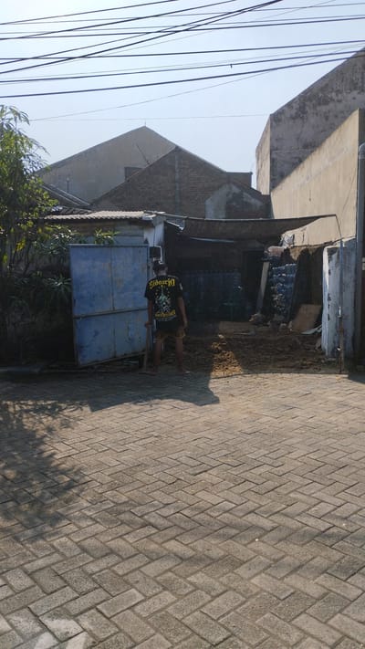 A man wearing a black t-shirt and shorts stands in front of a blue metal gate in the city of Tanjungsari, Indonesia. He holds a shovel in his right hand. Behind him, there are several buildings and stacks of plastic bottles. The ground is paved with interlocking bricks. Overhead, power lines crisscross the sky. The scene appears to be a residential or commercial area with some ongoing construction or renovation.