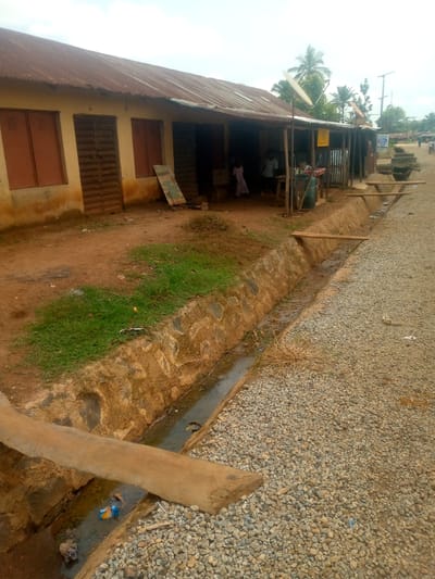 This image depicts a street scene in Madakiya, Nigeria, characterized by a small row of buildings with corrugated metal roofs. The buildings appear to be simple, possibly shops or residences, with wooden shutters on windows and doors. In front of the buildings, a narrow, rough trench runs parallel to the street, which is covered with gravel. Two wooden planks serve as makeshift bridges over the trench. A small amount of green grass grows on the bank of the trench closest to the buildings. The overall impression is of a modest, somewhat rural or semi-urban environment. The sky is overcast, suggesting a cloudy day. There are a few people visible in the scene, including a child in a light-colored dress standing near the entrance of one of the buildings and other indistinct figures further down the street. There are also some visible objects such as satellite dishes on the roofs, a blue barrel, and debris scattered along the trench and the roadside.