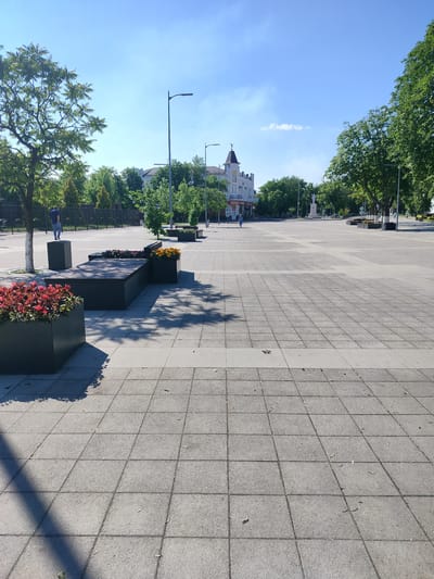 The image depicts a wide, paved public square in Labinsk, Russia, on a clear, sunny day. The square is laid with large, square concrete tiles arranged in a grid pattern. In the foreground, a dark, rectangular planter box is filled with vibrant red flowers. Adjacent to it is a long, dark, modular bench system, also featuring planters with red and orange flowers. Shadows from trees and the bench are cast across the tiled surface.
Further into the square, several lamp posts with modern designs are visible. In the mid-ground, a street with a few people walking can be seen, along with buildings lining the street. One building, with a distinctive red-tiled roof and white facade, appears to be an older architectural style. A statue on a pedestal stands in the distance. The background is filled with lush green trees and a bright blue sky with scattered white clouds. The overall impression is that of a well-maintained urban public space.