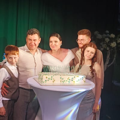A family of five is gathered around a cake on a table, with a dark green curtain in the background and white flowers to the right. The family consists of a man on the left with his arm around a young boy with red hair. Next to him is a woman in a white dress, and to her right is a man wearing glasses. On the far right is a young girl with braids. The cake is rectangular with white frosting decorated with green and gold leaves and has multiple green candles. The lighting suggests an indoor event, possibly a celebration. The location is identified as the city of Svetloe, Russia.