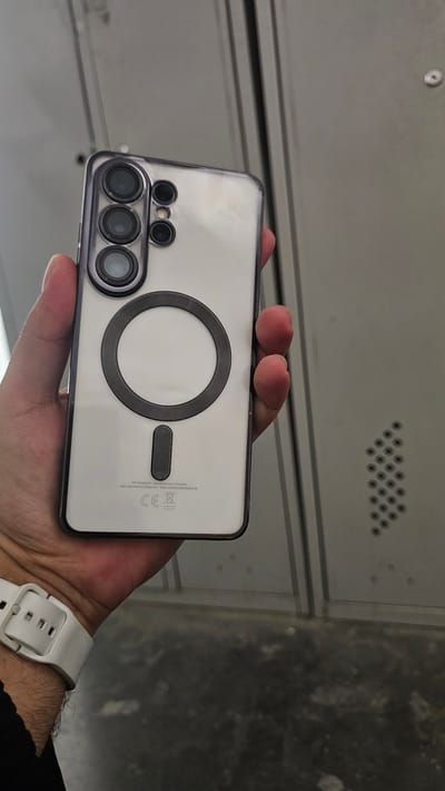 A person's hand, with visible wrist hair, is holding a white smartphone with a clear protective case. The back of the phone displays a circular magnetic charging ring with a smaller rectangular magnet below it. The camera module, featuring multiple lenses arranged in a vertical stack, is visible on the left side of the phone. The phone's casing has some subtle text and markings, including "SM-59488/DS Manufactured in Vietnam," "IMEI" numbers, and a "CE" logo. The background appears to be a metallic, textured surface, possibly lockers or shelving, with a row of small, dark circular perforations. The lighting suggests an indoor environment. No specific time of day or weather conditions can be determined.