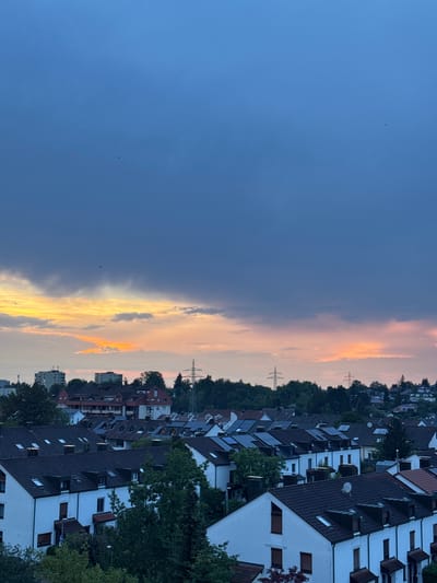 This is a photograph taken from an elevated perspective overlooking a residential area in Sindelfingen, Germany. The image captures a twilight sky with a dramatic gradient of colors, ranging from deep blue at the top to hues of orange and yellow near the horizon. Dark, heavy clouds dominate the upper half of the sky, while lighter, scattered clouds are visible closer to the sunset colors. Below the sky, a dense cluster of houses with dark tiled roofs fills the middle ground. The houses appear to be mostly two or three stories high and are characterized by white facades and numerous dormer windows. Solar panels are visible on the roofs of several houses, indicating a modern residential development. Green trees and foliage are interspersed among the buildings, and a few taller structures, possibly apartment buildings or businesses, are visible in the distance, along with utility poles and transmission towers. There are no people or obvious actions depicted in the scene. The overall impression is a quiet, suburban evening in Sindelfingen.