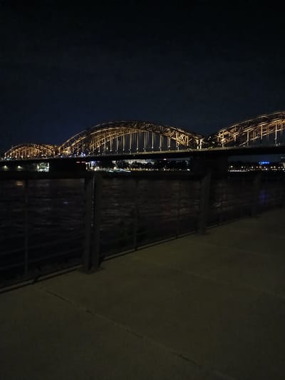 The image captures a nighttime view of the Hohenzollern Bridge in Cologne, Germany. The bridge, a prominent landmark, is illuminated with warm yellow lights, highlighting its intricate steel arches. Below the bridge, a dark river flows, reflecting the ambient light. A metal railing separates a paved walkway in the foreground from the river. The sky above is a deep, dark blue, typical of a clear night. The scene conveys a sense of urban tranquility and architectural grandeur after dark.