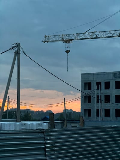 The image depicts a construction site at dusk in Froli, Russia. A tall yellow construction crane dominates the left side of the frame, its lattice structure silhouetted against a moody, overcast sky. The crane's boom extends diagonally upwards, with a hook mechanism visible at its end. In the background, a partially constructed concrete building with rows of dark, empty window frames stands out against the sky. A corrugated metal fence, likely part of the construction site's perimeter, runs vertically along the right edge of the image.
The sky displays a gradient of colors, transitioning from a deep, cloudy grey at the top to a warm, soft orange and pink hue near the horizon, indicative of sunset. This warm glow illuminates the distant cityscape, where the outlines of apartment buildings and trees are faintly visible. Power lines crisscross the scene, supported by a concrete utility pole in the foreground, adding to the urban industrial atmosphere.
The overall mood is one of quiet anticipation and the end of a day's work. The dimming light and the presence of the unfinished building suggest a pause in activity, with the construction project awaiting completion. There is no visible text in the image.