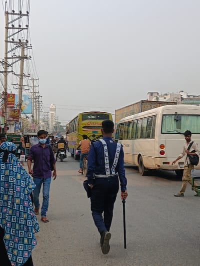 This image depicts an urban street scene in Savar, Bangladesh, under an overcast sky. The view is from a street-level perspective, featuring pedestrians and vehicles.
In the foreground, a police officer is observed from the rear, wearing a dark blue uniform with reflective "POLICE" text on shoulder straps and a reflective belt. The officer holds a black baton vertically in the right hand. To the officer's left, a person, also viewed from the rear, wears a blue and white patterned full-body garment and a head covering. Slightly forward and to the left of the police officer, a male individual walks towards the camera, dressed in a purple long-sleeved shirt and blue jeans, with a light blue surgical mask covering his mouth and nose.
On the right side of the mid-ground, a male pedestrian walks away from the camera, wearing a light brown collared shirt and khaki cargo pants, carrying a black backpack on his left shoulder.
The roadway ahead accommodates vehicular traffic. A yellow bus, displaying "HamEL" and Bengali script on its rear, is positioned on the left side of the road, facing away. Adjacent to it on the right, the rear section of a white mini-bus, marked "ACME" on its side, is also facing away. Behind the white mini-bus, a large, light-colored truck or container vehicle is partially visible. A motorcyclist and a person on a bicycle are present between the buses.
The left side of the street is lined with commercial buildings displaying various signs, including one with a green circular logo and Bengali text. Multiple utility poles with dense bundles of electrical and communication wires are prominent along this side. In the distant background, a white minaret-like structure rises against the light sky. The asphalt road surface appears dry.