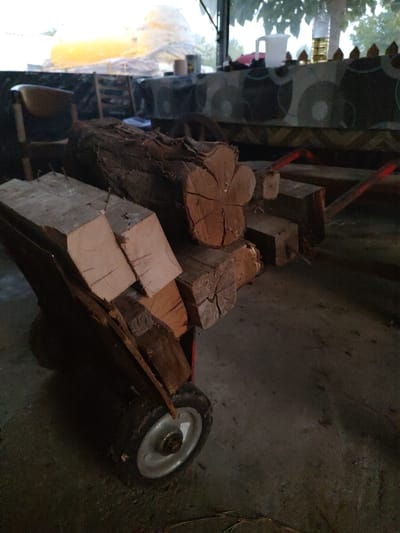 In Kochmar, Bulgaria, a close-up, low-angle shot captures a weathered wooden wheelbarrow loaded with cut logs. The wheelbarrow's metal rim and tire are visible on the right, partially obscured by the logs. The logs themselves appear to be of various sizes, some with rough bark and others sawn into rough rectangular shapes. The ground is a rough, concrete-like surface. In the background, out of focus, is a table covered with a patterned cloth, a white pitcher, and a bottle of oil. Beyond that, a large, blurry yellow object is visible, possibly an inflatable structure or tarp. Trees and foliage are visible through an opening in what appears to be a covered outdoor space. The overall lighting is dim, with some areas illuminated by what could be artificial light.