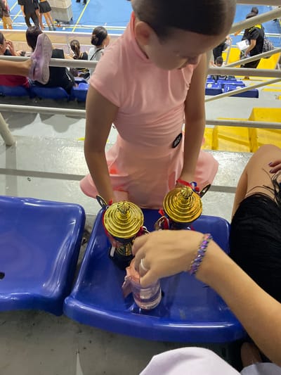 In a stadium with blue seats, two trophies are placed on a blue seat. A person in a pink dress is holding one of the trophies. Another person's arm, wearing a bracelet, is reaching for a small plastic cup next to the trophies. In the background, other people are seated, and a person in a black shirt with yellow accents is visible. The environment appears to be an indoor sports arena or event hall, possibly at a school or community center. There is no specific indication of the city of Thuận An, Vietnam within the image.