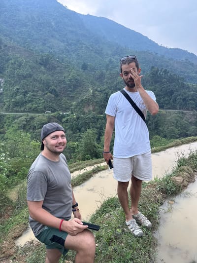 This image features two men situated in a mountainous, rural landscape within Nậm Dịch Commune, Vietnam. The scene showcases lush, forested mountains under a hazy or overcast sky, with what appear to be muddy rice paddies or irrigation channels bordering grassy banks in the foreground.
In the foreground, on the viewer's right, one man is visible. He wears a grey t-shirt, green shorts with black accents, and a grey cap. He has a beard and a subtle smile as he looks towards the left. He is holding a black smartphone, seemingly interacting with it. A red bracelet is on his right wrist, and a dark band, possibly a smartwatch, is on his left.
Further back and to the viewer's left, the second man is standing. He is dressed in a white t-shirt, light-colored shorts, and light grey sandals. Dark sunglasses rest on his head. With a beard and a slight smirk, he engages with the camera, making a playful gesture by partially covering his right eye with his hand. A black strap, likely from a bag, crosses his shoulder. He also holds a dark smartphone in his left hand.
The background consists of densely forested, steep mountains, disappearing into a soft haze. In the midground and foreground, the muddy water of the paddies or channels is visible, bordered by uneven ground with various grasses and small plants. The men's attire suggests casual outdoor activity or travel. The overall atmosphere conveys a relaxed moment during a daytime exploration of this scenic, agricultural mountain region, likely under diffused light from a cloudy sky. No discernible text is visible on any objects or clothing.