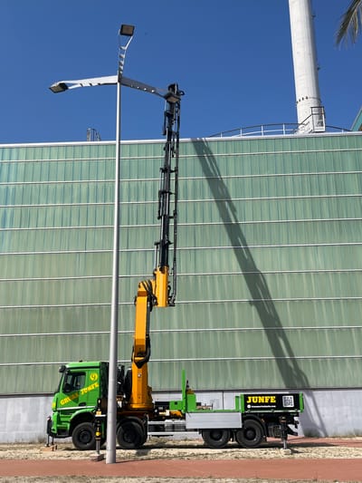 A bright green truck with a yellow crane is parked on a sandy surface with a red-colored path in the foreground. The truck is branded with "GRÚAS JUNFE" on the side of the cab and "JUNFE" with a QR code and website address on the rear. The crane is extended vertically, casting a long shadow on the glass facade of a modern building behind it. The building has horizontal green glass panels and is adjacent to a tall, white cylindrical structure, possibly a chimney or industrial tower. A street lamp with multiple lights is also visible to the left of the truck. The sky is a clear, vibrant blue. The location is identified as Sant Adrià de Besòs, Spain, likely near an industrial or commercial area. There are no people visible in the image.