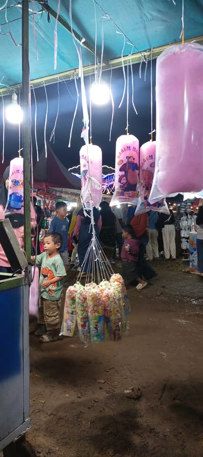 In Sinar Semendo, Indonesia, an outdoor night market stall is depicted. The stall is covered by a blue tarp supported by metal poles. Hanging from the tarp are several large, clear plastic bags filled with pink cotton candy, some featuring cartoon characters. Below the cotton candy, a cluster of smaller plastic bags, also containing colorful snacks (likely popcorn), are suspended from strings. A young boy, wearing a green t-shirt with a dinosaur graphic and beige cargo shorts, stands in front of a counter-like structure on the left, appearing to interact with a display. Other individuals, including adults and children, are visible in the background, some browsing at other stalls or walking through the muddy ground. The lighting appears to be artificial, with bright bulbs illuminating the scene. The ground is covered in dirt and has some puddles, suggesting it may have recently rained or is an unpaved area. The overall atmosphere is that of a local, casual fair or market.