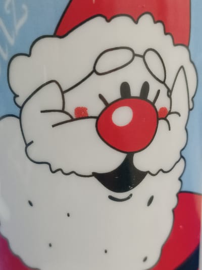 The media file is a close-up, detailed shot of a cartoon depiction of Santa Claus. The subject is the face of Santa, featuring his iconic red hat, white beard, round red nose with a white highlight, and round glasses. Two small red circles are visible on his cheeks, representing rosy cheeks. The background is a light blue with faint white, curly, abstract shapes, possibly representing snow or a festive pattern. The overall tone is cheerful and festive, characteristic of holiday imagery. There is no discernible text within the visible portion of the image. The setting and time of day are not specified beyond the implied context of a holiday illustration.